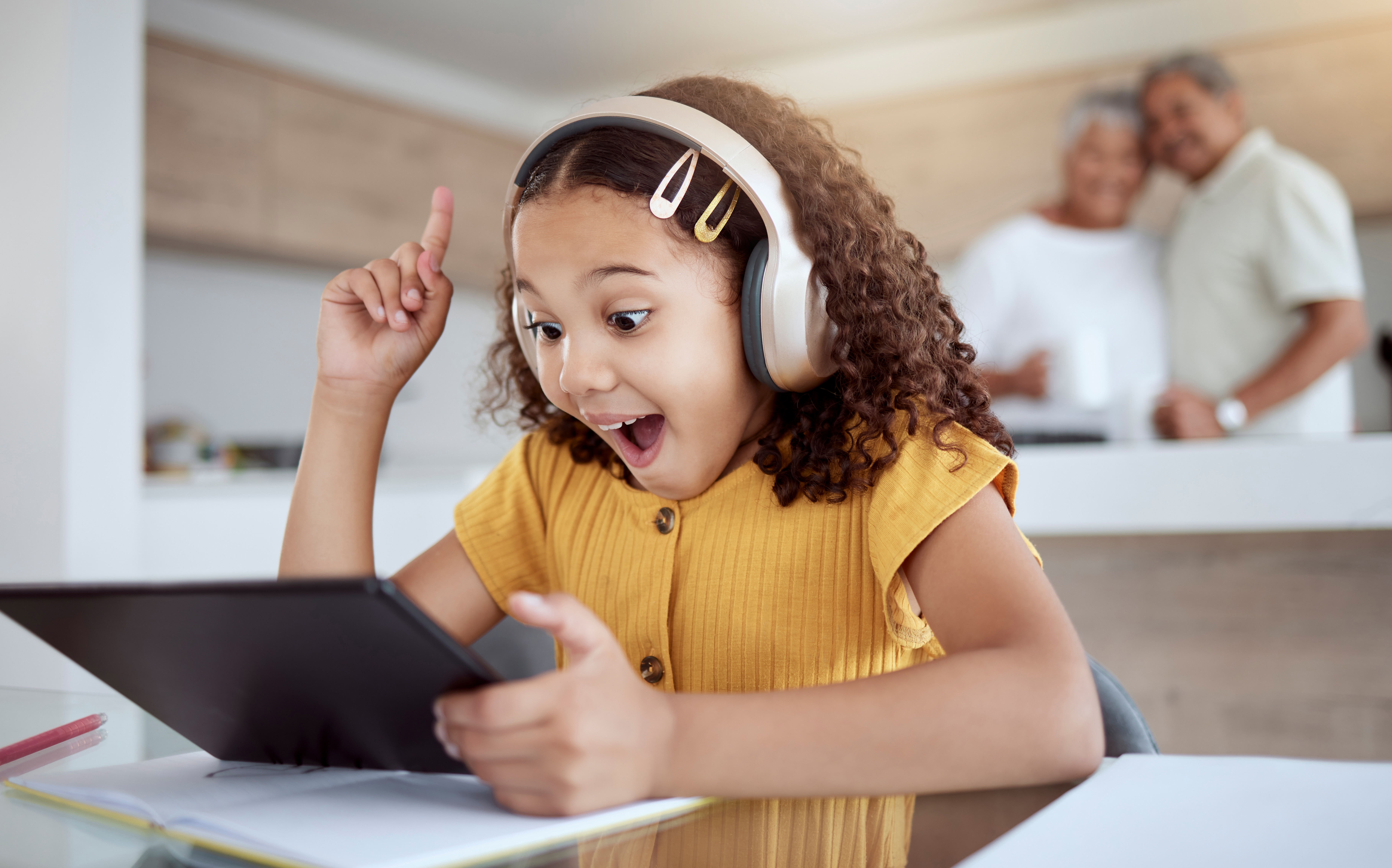 Excitedly, an elementary-aged, Latina girl wearing headphones listens to a podcast episode on her tablet while seated at her kitchen table. Standing behind the kitchen island, her grandparents stand with an arm around each other and smile.