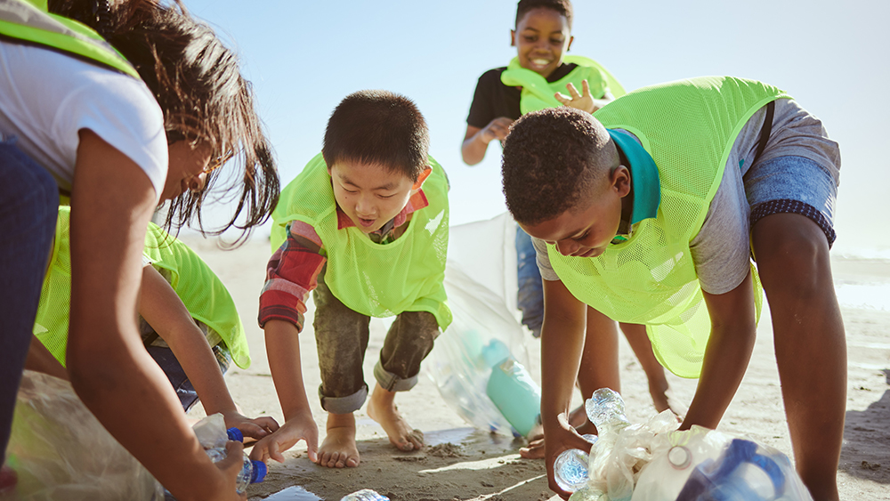 Four grade-school students, wearing neon yellow pinnies, pick plastic bottles off the sand at the beach.