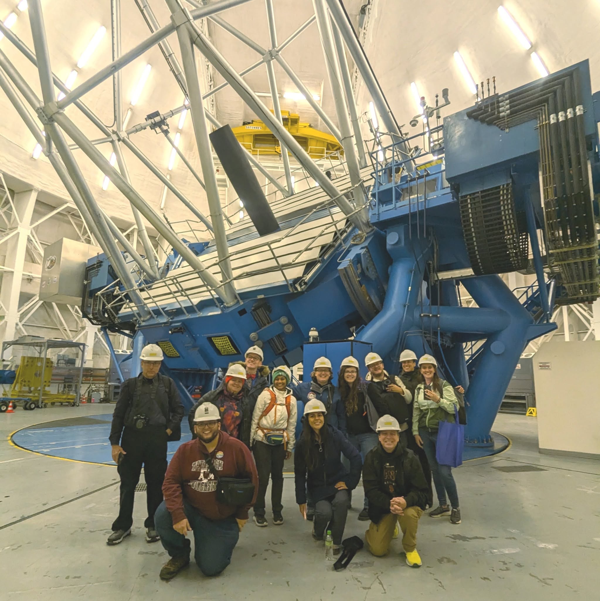 A group of people wearing hard hats are posing in front of a large telescope inside an observatory. The telescope is prominently displayed with its intricate structure and blue coloring. The group took part in a an educational visit.