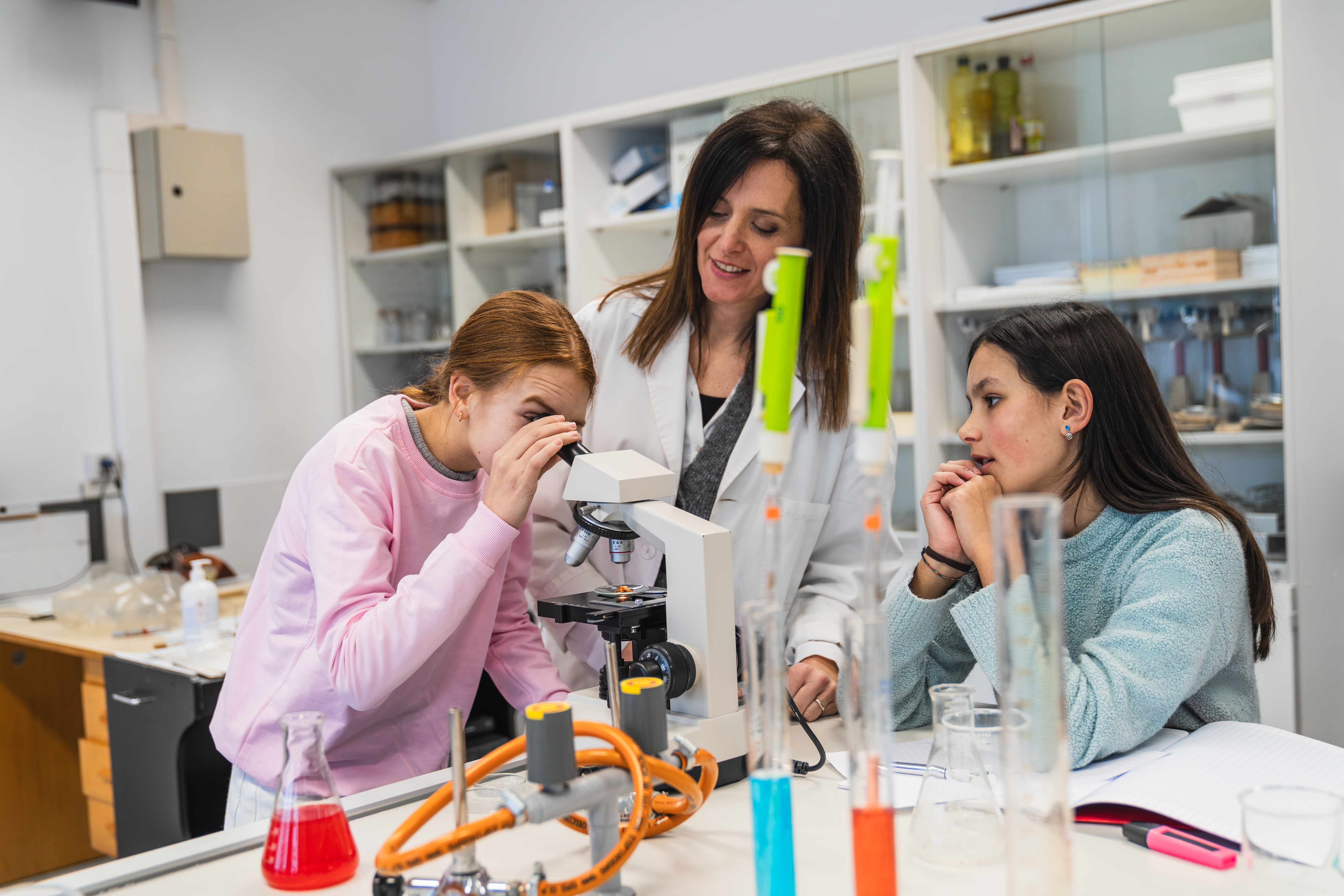 Three individuals are in a laboratory, an adult and two junior high students, engaged in a science experiment. One student is looking through a microscope, while another watches. The adult smiles, standing nearby. The table is filled with lab equipment, including test tubes with colored liquids and a coiled hose. Shelves stocked with bottles and containers are in the background.