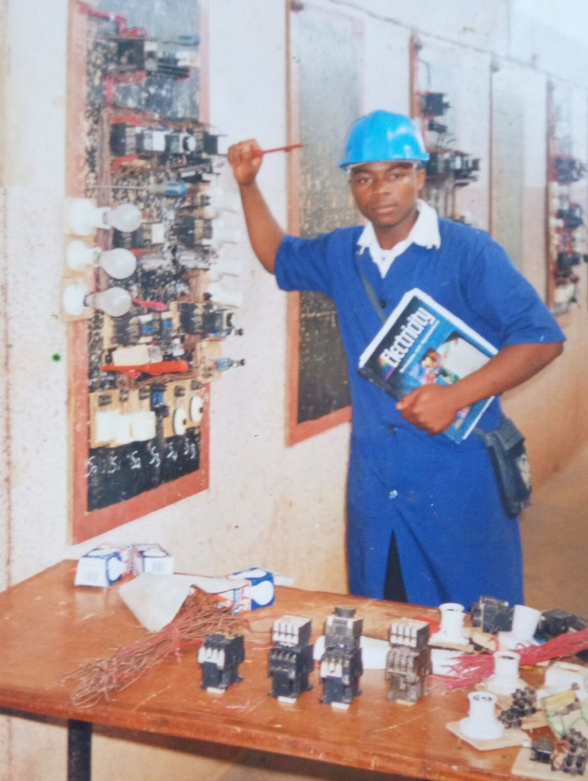 Desmond Wysenyuy in a blue uniform and hard hat holds a textbook while standing next to a wall with electrical panels. A table in the foreground displays various electrical components.
